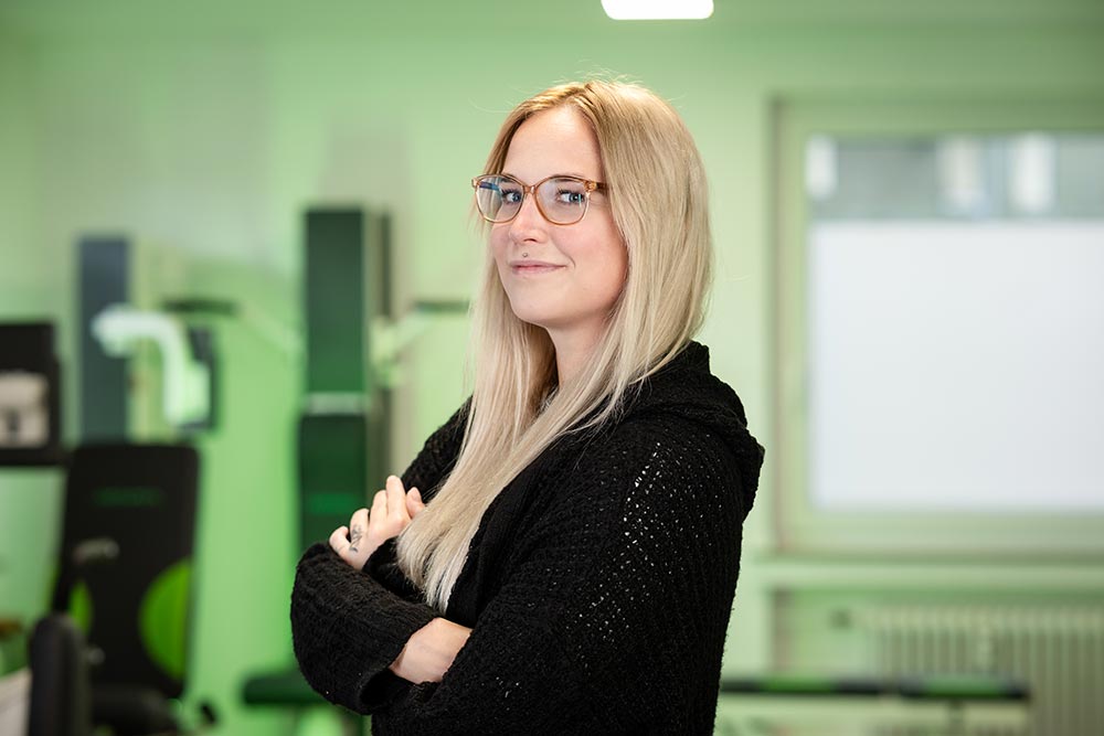 Blonde Frau mit Brille, neutraler Blick, schwarzer Pullover, Laborgruen