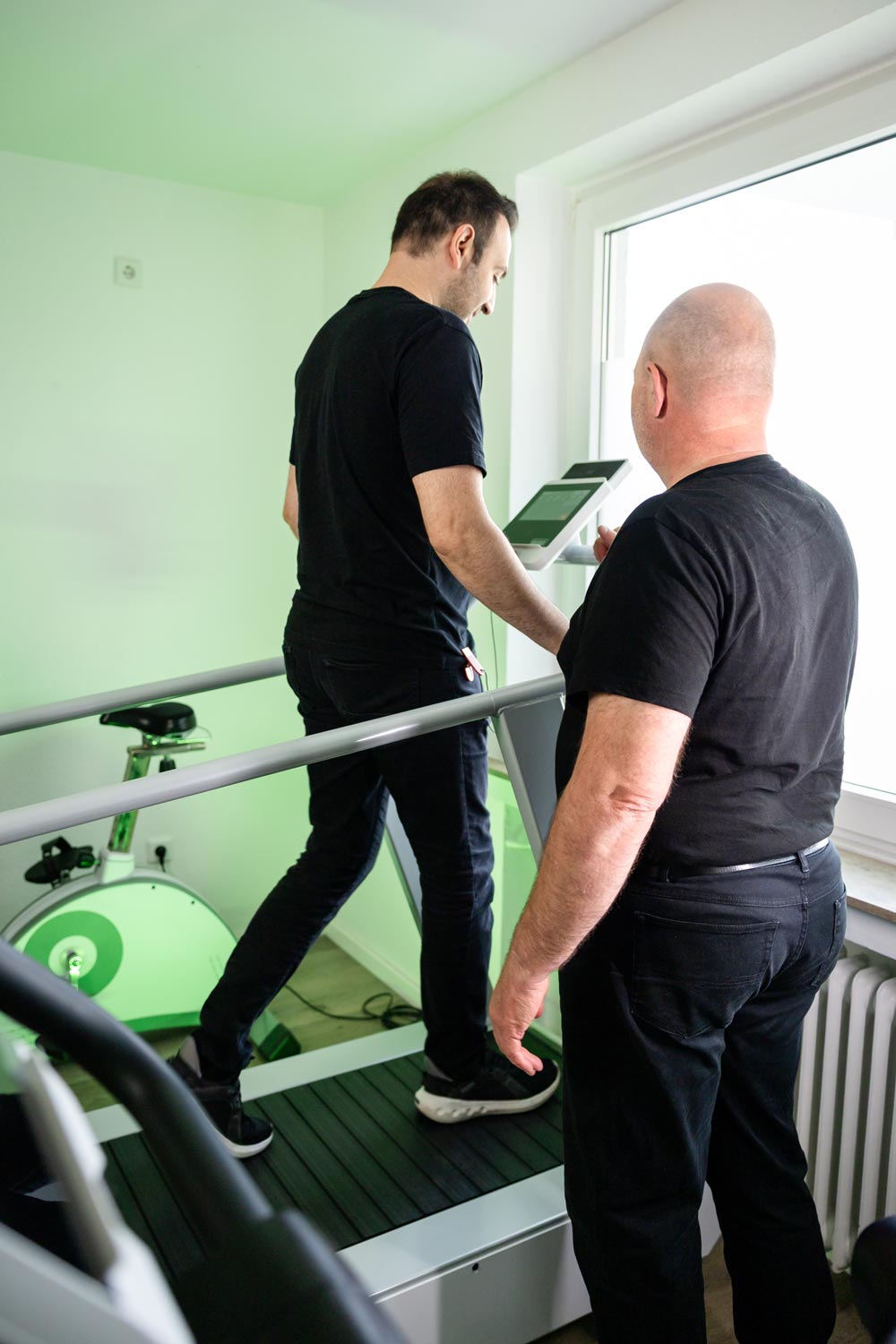 Zwei Maenner in Schwarz, konzentriert, am Laufband mit Display, gruenes Rad hinten
