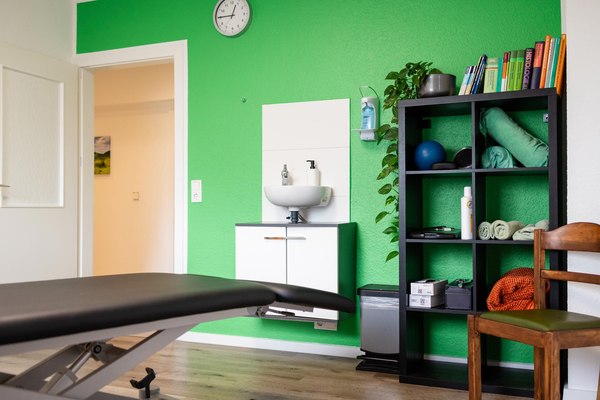 Leerer Therapieraum, ruhig, grüne Wand, Regal mit Matten und Ball, Liege links