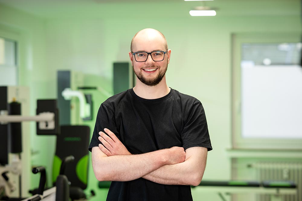 Glatzkopf mit neutralem Blick im schwarzen Shirt, Arme verschraenkt, gruenes Fitnessstudio